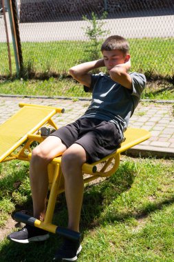 Genç bir adam açık hava fitness ekipmanlarında mekik çekiyor. Spor kıyafetleri giyiyor, güneşli, çimenli bir park ortamında antrenmanına odaklanıyor..