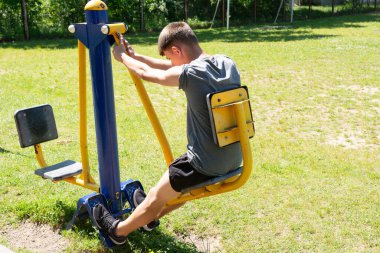 Genç bir insan açık hava spor salonunda bacak pres makinesi kullanıyor. Güneşli gün ve yeşil çimenler sağlıklı yaşam tarzını ön plana çıkarıyor..