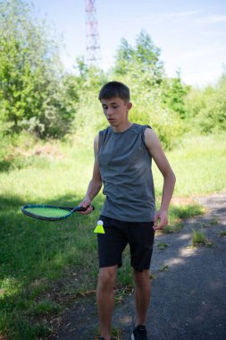 Genç bir adam parka benzer bir ortamda badminton oynamaktan hoşlanır. Atletik kıyafetler giyiyor ve raket tutuyor. Bir sonraki servis için hazır..