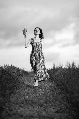 Bulutlu bir günde, elinde ayakkabılarıyla lavanta tarlasında çıplak ayakla yürüyen çiçekli bir kadın resmi..