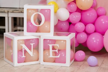 Üç şeffaf küp kutu, 'One' yazılıyor, pembe ve şeftali balonlarıyla dolu. İlk doğum günü partisi dekorasyonu ya da fotoshoot desteği için ideal.