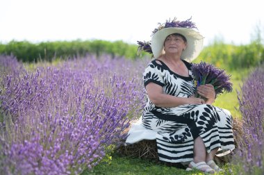 Siyah-beyaz elbiseli ve geniş kenarlı şapkalı bir kadının sakin bir sahnesi, canlı bir lavanta tarlasında saman balyasında oturuyor, elinde bir demet lavanta tutuyor..