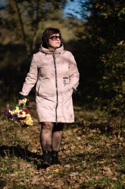 Pembe paltolu bir kadın, canlı bir buket taşıyan, güneş lekeli bir orman yolunda yürüyor. Güneş gözlüğü ve siyah çizme giyiyor. Huzurlu bir doğa sahnesi..