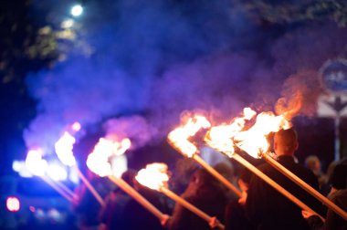 Birkaç birey karanlıkta yürüyor, her biri havada yanan bir meşale tutuyor. Alevler sırtlarını aydınlatıyor ve gece gökyüzüne karşı çarpıcı bir görsel zıtlık oluşturuyor..