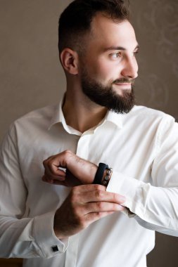 Kol düğmelerini ve kol saatini titizlikle ayarlayan beyaz gömlekli bir adamın portresi. Sakalı ve kısa saçı var. Temiz ve sofistike bir tarzı var..