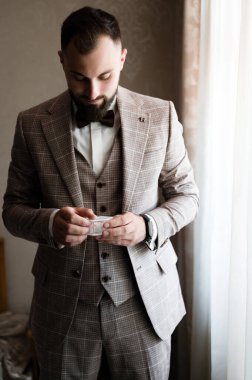 Süslü kahverengi bir takım elbise ve papyon giymiş bir damat, dikkatlice küçük bir kutu tutuyor. Pencerenin yanında duruyor, beklenti ve ciddiyet havası yayıyor..