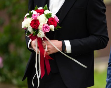 Siyah takım elbiseli bir damadın canlı bir düğün buketini nazikçe tutuşu. Çiçekler kırmızı, pembe ve beyaz güllerin karışımı, zarif kurdelelerle bağlanmış..