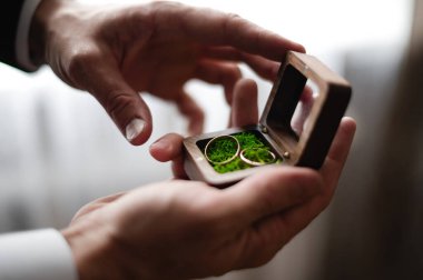 A person delicately holds a small, wooden box containing two gold wedding bands. The rings rest on a bed of green moss, adding a touch of nature to the scene. clipart