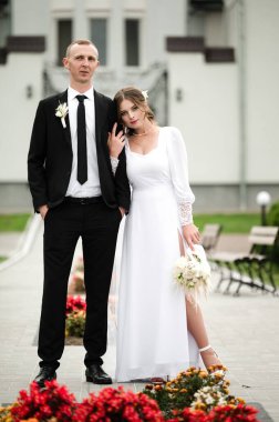 Şık bir gelin ve damat, düğün kıyafetlerini sergilemek için birbirlerine yakın dururlar. Fotoğraf dışarıda, ön planda canlı çiçeklerle çekiliyor..