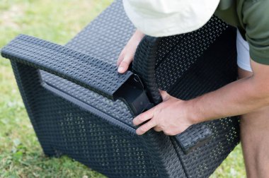 Bir insan açık havada koyu renkli hasır bir sandalye yapar. Sandalye örülmüş plastikten yapılmış ve sağlam görünüyor. Her yer çimenlik..