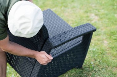 Koyu renkli hasır bir sandalye monte eden birinin yakın çekimi. Sandalye dışarıda çimenli bir çimenlikte birleştiriliyor..