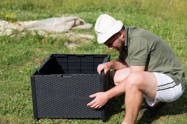 Bir adam çimenli bir alanda koyu gri plastik bir bahçe yatağı kuruyor. Çömeliyor, parçaları dikkatlice birleştiriyor. Hava güneşli ve açık..