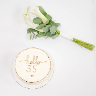 A celebratory scene featuring a simple white cake decorated with the words Hello 35 in gold. A small bouquet of white calla lilies and eucalyptus rests beside it. clipart