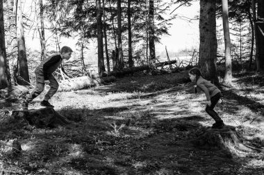 İki çocuğun siyah beyaz fotoğrafı, bir oğlan ve bir kız, bir orman ortamındaki ağaç kütüklerinden neşeyle atlıyorlar. Güneş ışığı ağaçların arasından süzülür.