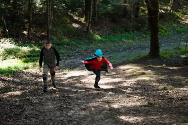 İki çocuk, ağaçların arasında yaratıcı oyunlar oynayarak güneşli bir günün tadını çıkarıyor. Kapüşonlu pelerinli bir çocuk öne fırlıyor. Orman sahnesini dolduran bir macera duygusu.