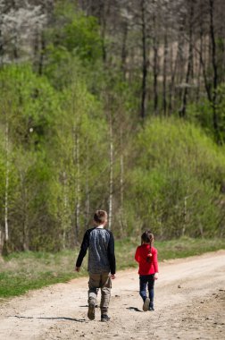 İki çocuk, bir oğlan ve bir kız, kırsal bir yolda geziniyorlar. Arka planda canlı bir bahar ormanı görülüyor. Huzurlu ve doğal bir ortam yaratıyor..