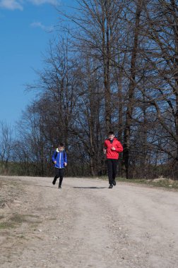 Bir çift bireyin açık mavi gökyüzünün altında yapraksız ağaçlarla çevrili pis bir yolda koştuğu görülüyor. Sağlıklı bir açık hava aktivitesinden hoşlanıyor gibiler..