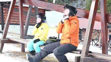 Bir adam ve bir çocuk, kayak kıyafetleri içinde, tahta bir bankta oturuyorlar. Kasklarını ayarlıyorlar, yamaçlarda bir güne hazırlanıyorlar. Arka planda karlı manzara görülebilir..