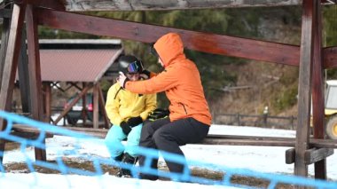 Karlı bir açık havada kayak kasklarıyla çocuklarına yardım eden bir ebeveynin yürek ısıtan bir sahnesi. Tahta bir bankta oturuyorlar, kış sporlarına hazırlar..