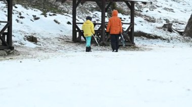 Kış sporu kıyafeti giymiş iki kişi kayak taşır ve kırsal bir ahşap barınaktan uzaklaşır. Zemin taze karla kaplı, sakin bir kış manzarası yaratıyor..