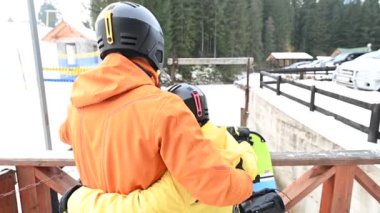 İki snowboardcu, kask ve parlak ceketler giyiyorlar, park yerine bakan karlı bir köprüde duruyorlar. Snowboard yapmaya devam etmeden önce dinleniyor gibi görünüyorlar..