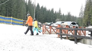 İki snowboardcu, bir yetişkin ve bir çocuk tahta bir çitin yanında karlı bir patika boyunca yürüyorlar. Park halindeki arabalar arka planda görülebilir, kozalaklı bir ormanın içinde..