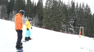Bir snowboard eğitmeni bir öğrenciye hafif bir yamaçtan aşağı doğru rehberlik eder. Karlı sahne, yemyeşil ağaçların olduğu bir zemin üzerine kurulmuştur. Snowboard öğrenmek için bir kış harikalar diyarı.