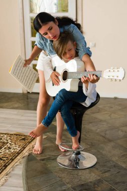 Bir kadın sabırla oğluna beyaz akustik gitar eğitimi veriyor. Bir tabureye oturup müziğe odaklanıyorlar. Sıcak, evcil bir öğrenme anı.