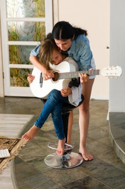Beyaz akustik gitar çalmayı öğrenen bir kadın, çocuğunun ellerine nazikçe rehberlik eder. Ev gibi, samimi ve sıcak..
