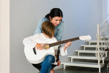 Küçük bir çocuk bir taburede oturur ve bir yetişkinden gitar dersi alır. Bir ev ortamında sağlanan enstrüman ve talimatlara dikkatle odaklanırlar.
