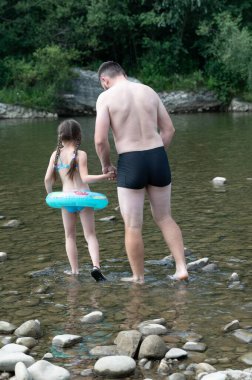 Bir adam ve genç bir kız, el ele, sığ ve kayalık bir nehirde dikkatlice yürüyorlar. Kızın küçük şişirilebilir bir yüzüğü var. Yemyeşil yapraklar nehir kenarlarında..