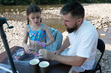 Bir adam ve genç bir kız nehir kenarındaki bir masada oturuyor, küçük renkli boncuklar ve diğer malzemelerle el işi yapıyorlar. Hareketlerine odaklanmış ve meşgul görünüyorlar..