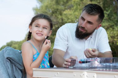Babası dışarıda güneşli bir günün tadını çıkarırken genç bir kız makyaj yapıyor. Bir masada oturuyorlar, piknik ya da açık hava oyun saati öneriyorlar..