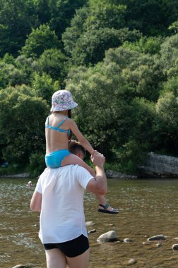 Sakin bir nehrin kenarında babasının omuzlarında mayo giymiş genç bir kız oturuyor. Bu cennet gibi yaz manzarası için yemyeşil ağaçlar bir zemin oluşturur. Suyun kenarında bir günün tadını çıkarıyorlar..