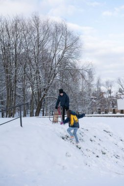Karla kaplı bir parkta iki çocuk oynuyor. Bir çocuk kızağını çekerken diğeri hafif bir yamaçtan aşağı koşuyor. Arkaplanda çıplak ağaçlar ve parçalı bulutlu bir gökyüzü var..