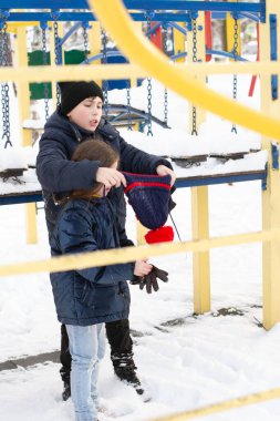 İki çocuk, bir erkek ve bir kız, karla kaplı bir oyun alanındalar. Çocuk kız kardeşinin bir şapka takmasına yardım ediyor. Sıcak kışlık giysiler giymişler..