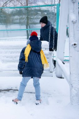 Kış kıyafetleri içinde iki çocuk, karlı bir alanda açık havada oynuyorlar. Arka planda beyaz bir salıncak seti görünür.