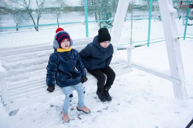 Sıcak şapkalar, ceketler ve botlar giymiş iki genç çocuk, karlı kış manzarasında beyaz bir ahşap salıncakta sessizce oturuyorlar. Huzurlu ve huzurlu bir sahne..