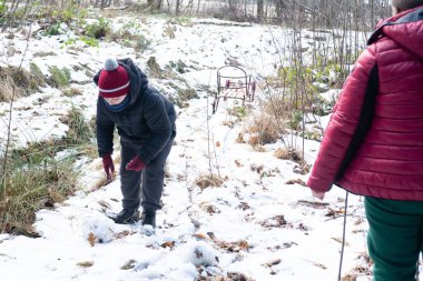Kışlık giysiler içindeki bir çocuk ağaçlık bir alanda karla kaplı bir yolda dikkatlice bir kartopu şekillendirir. Yakınlarda eski bir kızak var. Arka planda bir kişi daha görünüyor.