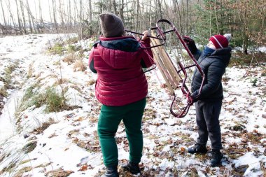 İki birey, bir yetişkin ve bir çocuk, ormandaki karlı bir yolda ilerliyorlar. Eski bir ahşap kızağı dikkatle taşıyorlar, kış gezintisinin tadını çıkarıyorlar..