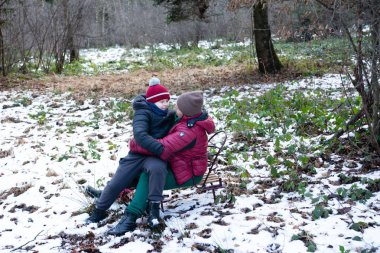 Bir ebeveyn ve çocuk, hafif karla kaplı bir ormanın ortasında kızağın üstünde hassas bir anı paylaşıyor. Çocuk yuvasına yakın, kış harikalar diyarının tadını çıkarıyor..