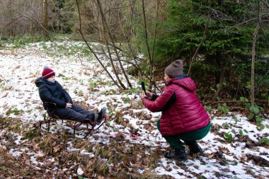 Küçük bir çocuk kışın ormanlık alanda küçük bir kızağın üzerinde oturur. Yakınlarda bir yetişkin, telefonuyla fotoğraf çekiyor..