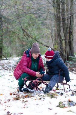 Bir yetişkin ve küçük bir çocuk, küçük bir ahşap kızağın yanında karda oturup akıllı telefona bakıyorlar. Kışlık giysiler giymişler..