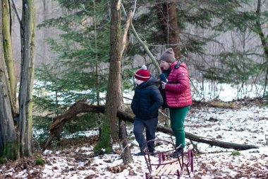 Karla kaplı bir ormanda bir kadın ve çocuk yürüyor. Çocuk kışlık kıyafetlerle sarılı. Yakınlarda bir ahşap kızak var. Kış eğlencesi olduğunu ima ediyor..