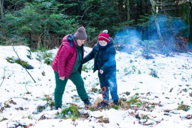 Bir yetişkin ve bir çocuk karlı bir ormanda küçük bir ateşe bakıyor. Açık hava macerasının tadını çıkarıyor gibi görünüyorlar. Sahne huzurlu ve bir sıcaklık hissi uyandırıyor..