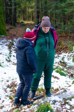Sıcak giyimli bir yetişkin ve çocuk, karlı bir kış ormanında küçük bir ateşin yanında dururlar. Ateşe eğilimli görünüyorlar, doğada sakin bir anın tadını çıkarıyorlar..