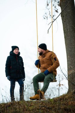 Bir adam ve bir çocuk, ağaçta sallanan salıncakta birlikte oturarak huzurlu bir kış gününün tadını çıkarıyorlar. Onlar kışlık giysiler giydiler. Arka plan karlı bir manzara..