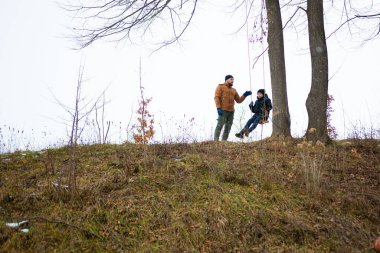 Bir adam çocuğunun ağaçta sallanışını izliyor. Sahne kışın bir parkta ya da ormanlık alanda geçiyor. Toprak kurumuş yapraklarla kaplı..