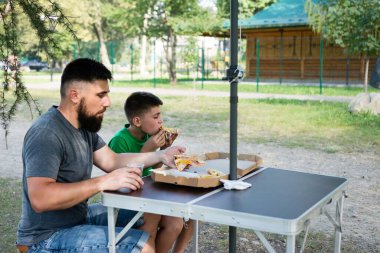 Sakallı bir adam ve küçük oğlu parkta portatif bir masada oturup pizza dilimlerini paylaşıyorlar. Kısmi bir ağacın gölgesinde gündelik bir yemeğin tadını çıkarıyorlar..