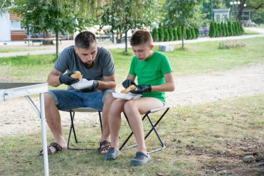 Bir adam ve bir çocuk açık havada küçük bir masada oturup tek kullanımlık konteynırlardan yiyorlar. Günlük kıyafetler giyiyorlar ve rahat görünüyorlar..